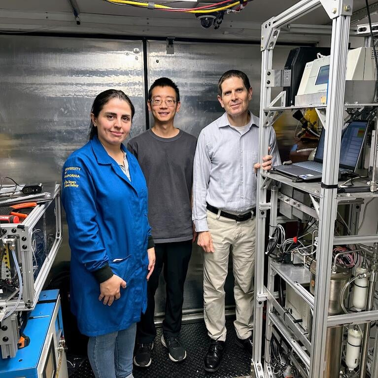 A front-view, full-body shot of three UCR CE-CERT researchers standing inside a mobile lab truck. From left to right: graduate student Maryam Yousefi, in a blue university jacket; graduate student Minghao Han, in a gray t-shirt and glasses; and CE-CERT Director Dr. Don Collins, in a collared shirt. They are all looking at the camera and smiling faintly. The mobile lab has shiny, corrugated metal walls and is filled with intricate scientific equipment on metal racks, with many wires visible.
