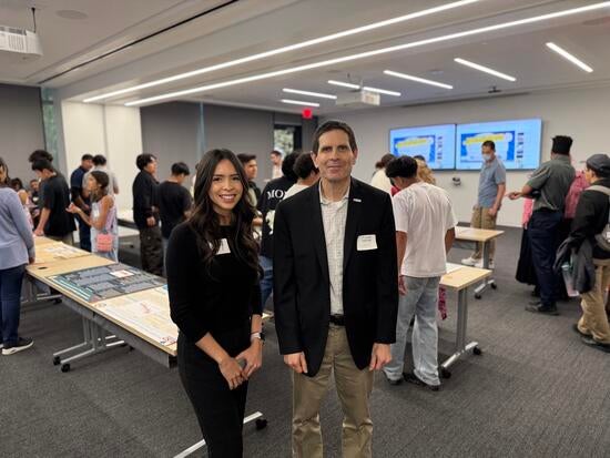 Kiara Salgado, a young woman with long dark hair wearing a black outfit, stands next to Don Collins, who is wearing a black blazer and khaki pants. They are both smiling at the camera in a classroom filled with students and poster presentations displayed on tables in the background.
