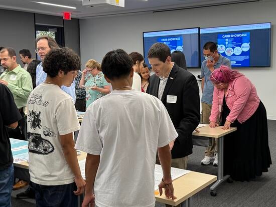 A group of students and adults are gathered in a classroom setting. Don Collins, wearing a black blazer and name tag, is standing at a table and reading a student’s project display. Other attendees are observing posters and engaging in conversations around the room.
