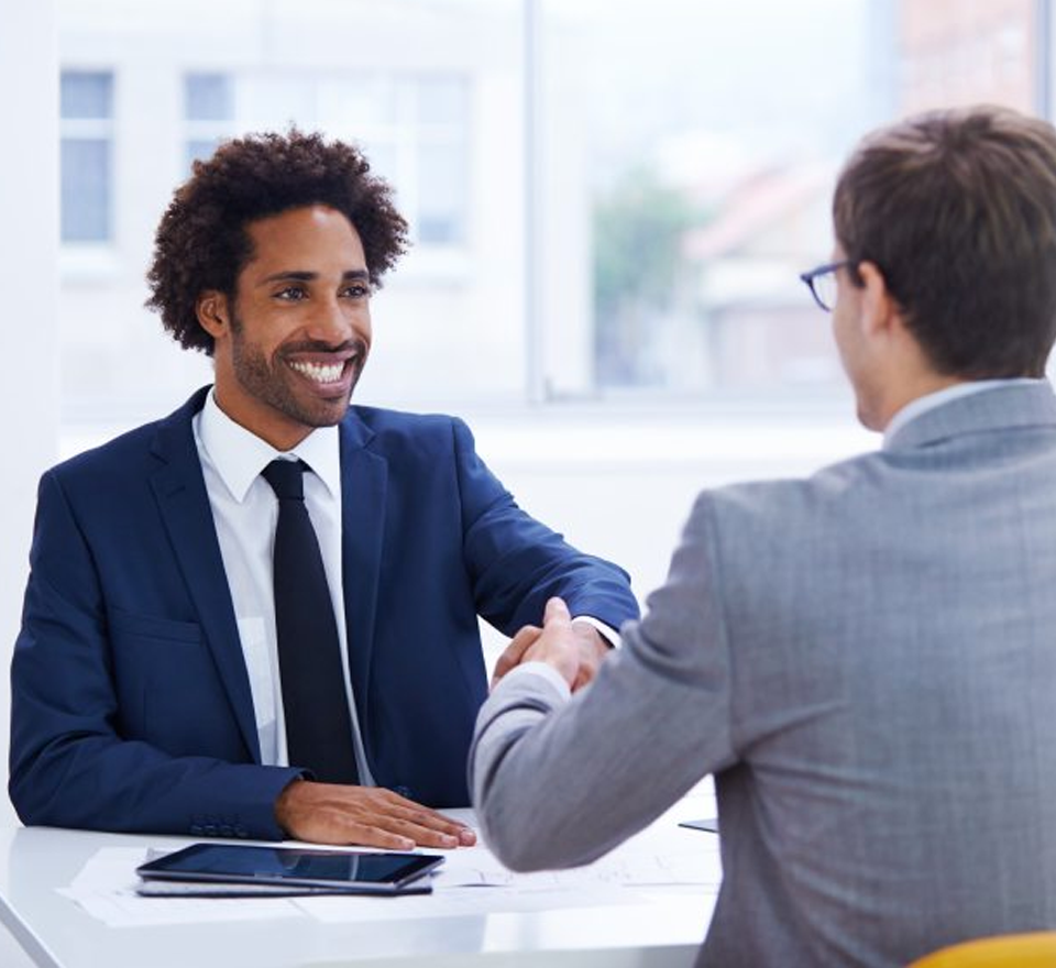 Young Black man shaking hands across a desk with a young White man in an office Both men in business suits