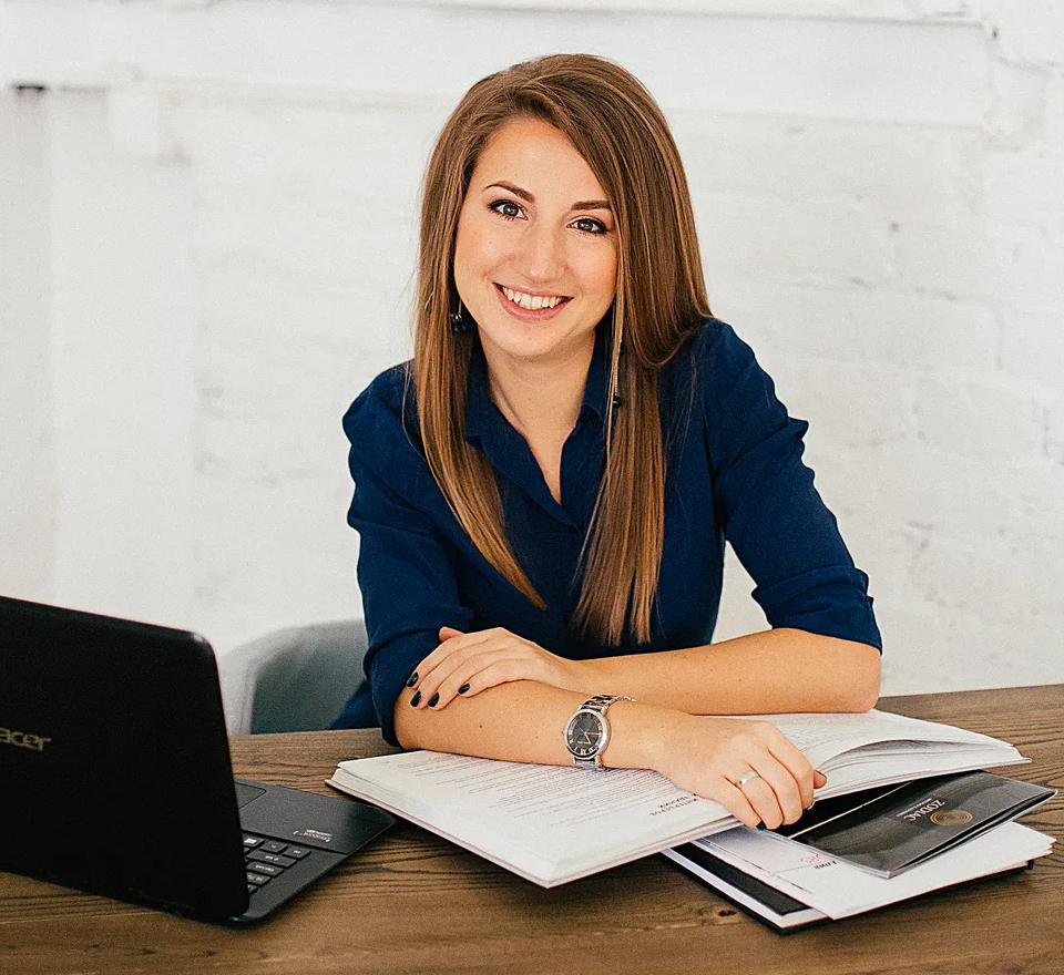 Young White business woman smiling at her desk with an open book under her arm and a laptop on the desk