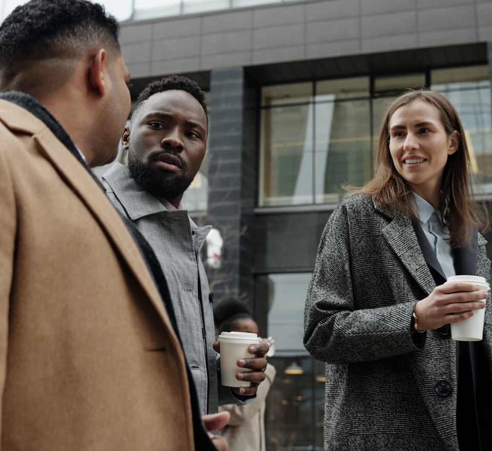 Two young men and a young woman wearing coats and holding coffee cups while talking and walking down a city street