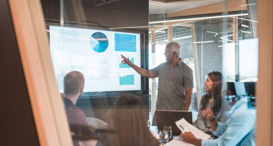 Older man giving presentation with onscreen graphs in a meeting