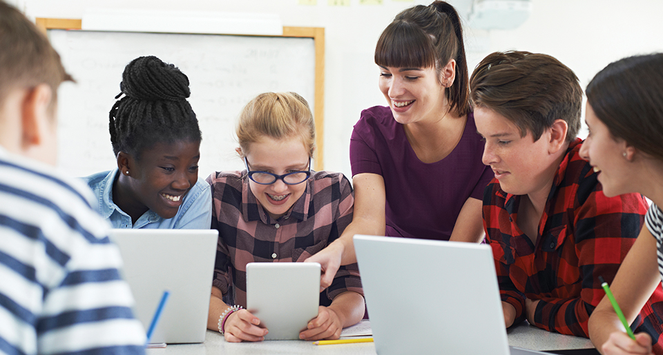 Diverse group of middle school students with laptops and tablets at a table with a teachers hand pointing at one of the screens