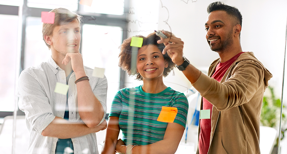Two young business men and young business woman smiling and looking at a glass board with post-its