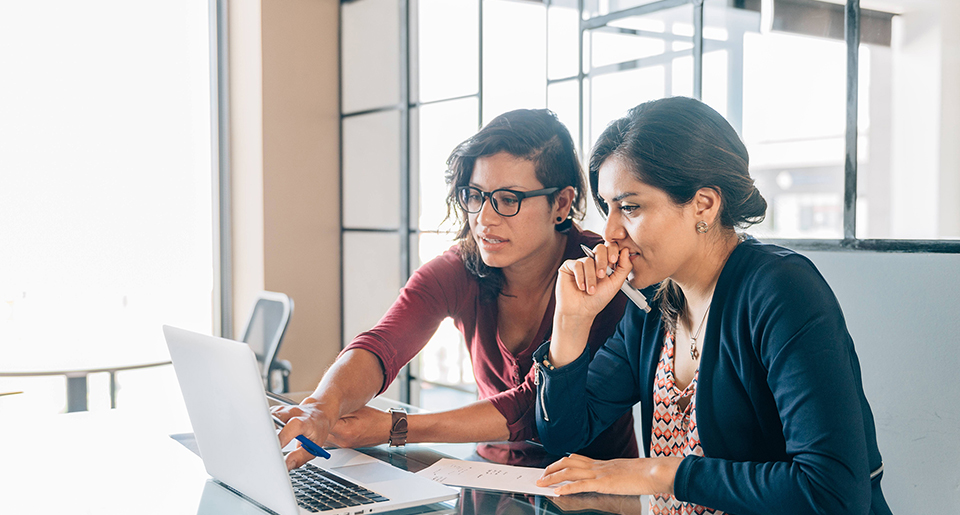 Two young Hispanic women one wearing glasses sitting in front of a monitor pointing at it